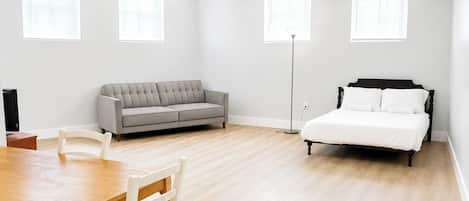 A room with a wooden table, a grey sofa, a white bed, and a floor lamp.