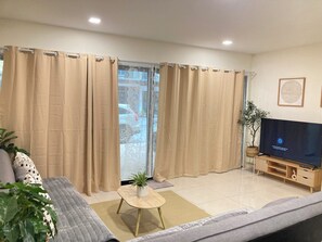 A living room with beige curtains, a sofa, a coffee table, a TV on a wooden stand, and a plant. - 4BR Townhome  Sleeps 10  Near Nimman & MAYA mall (Chang Wat Chiang Mai)