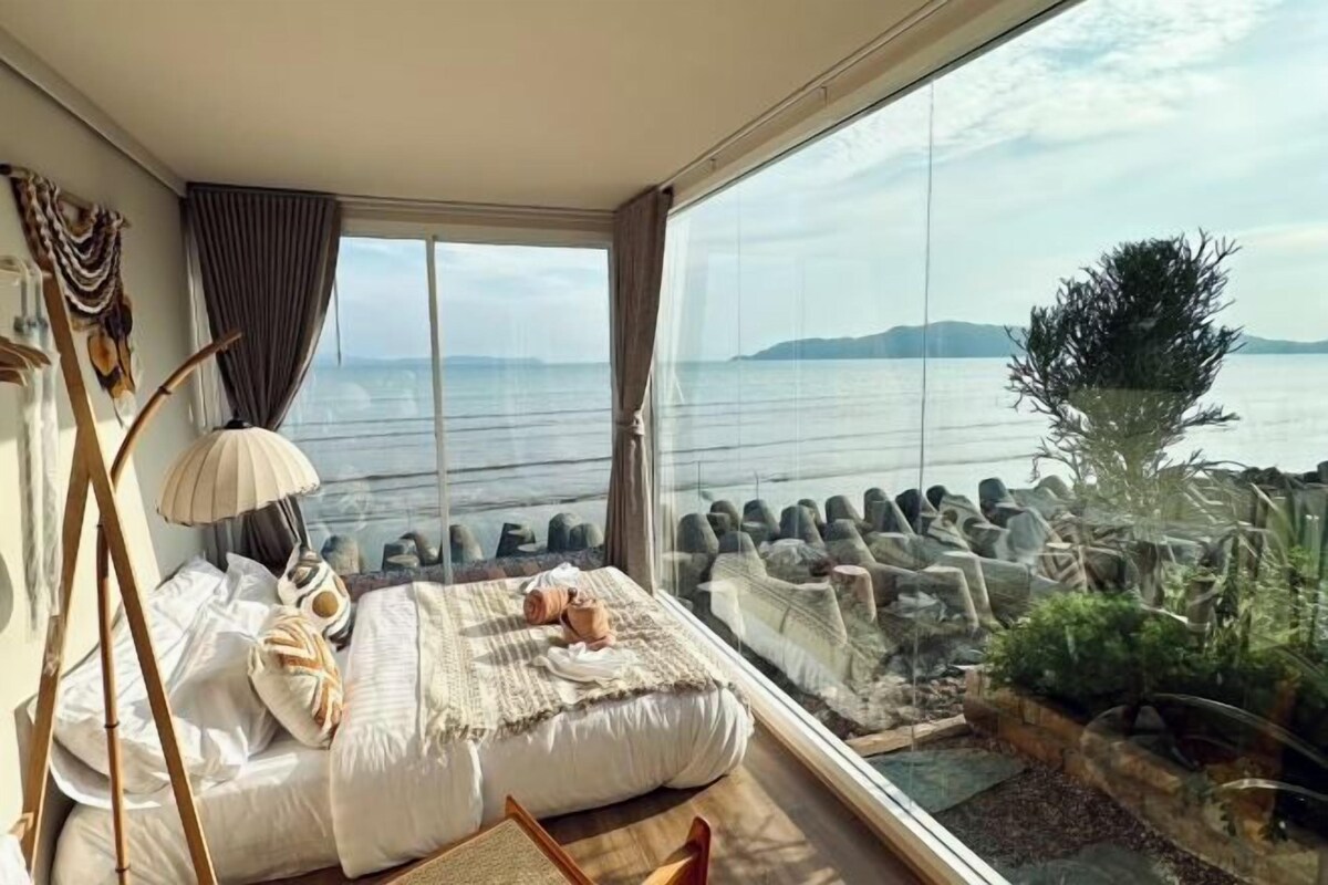 A bedroom with a large window, a bed, a lamp, and a view of the sea.