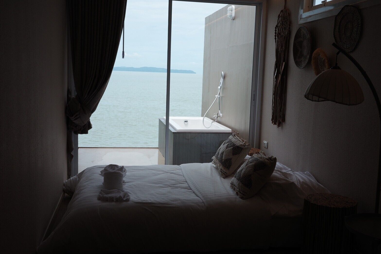 A hotel room with a bed, a window with a view of the sea, a bathtub, and a wall-mounted lamp.