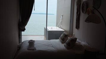 A hotel room with a bed, a window with a view of the sea, a bathtub, and a wall-mounted lamp.