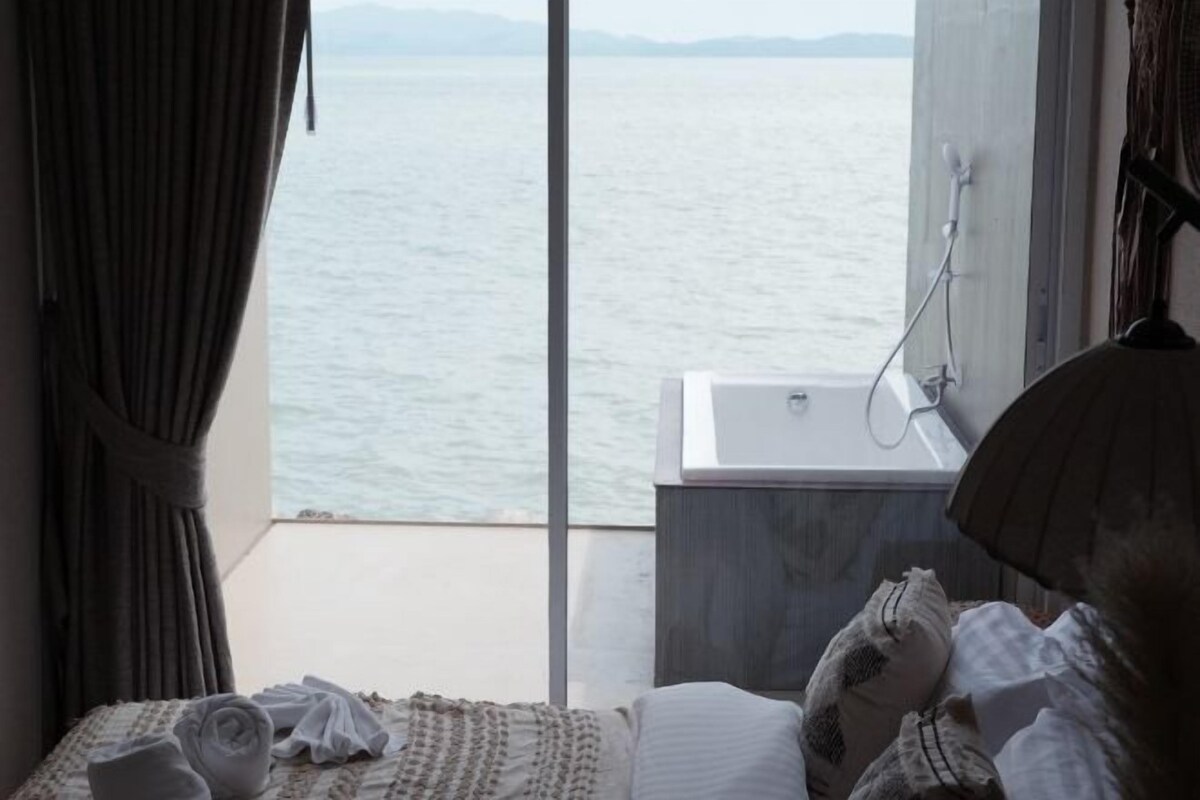 A room with a bed, a window with a view of the water, a sink, and a towel on the bed.