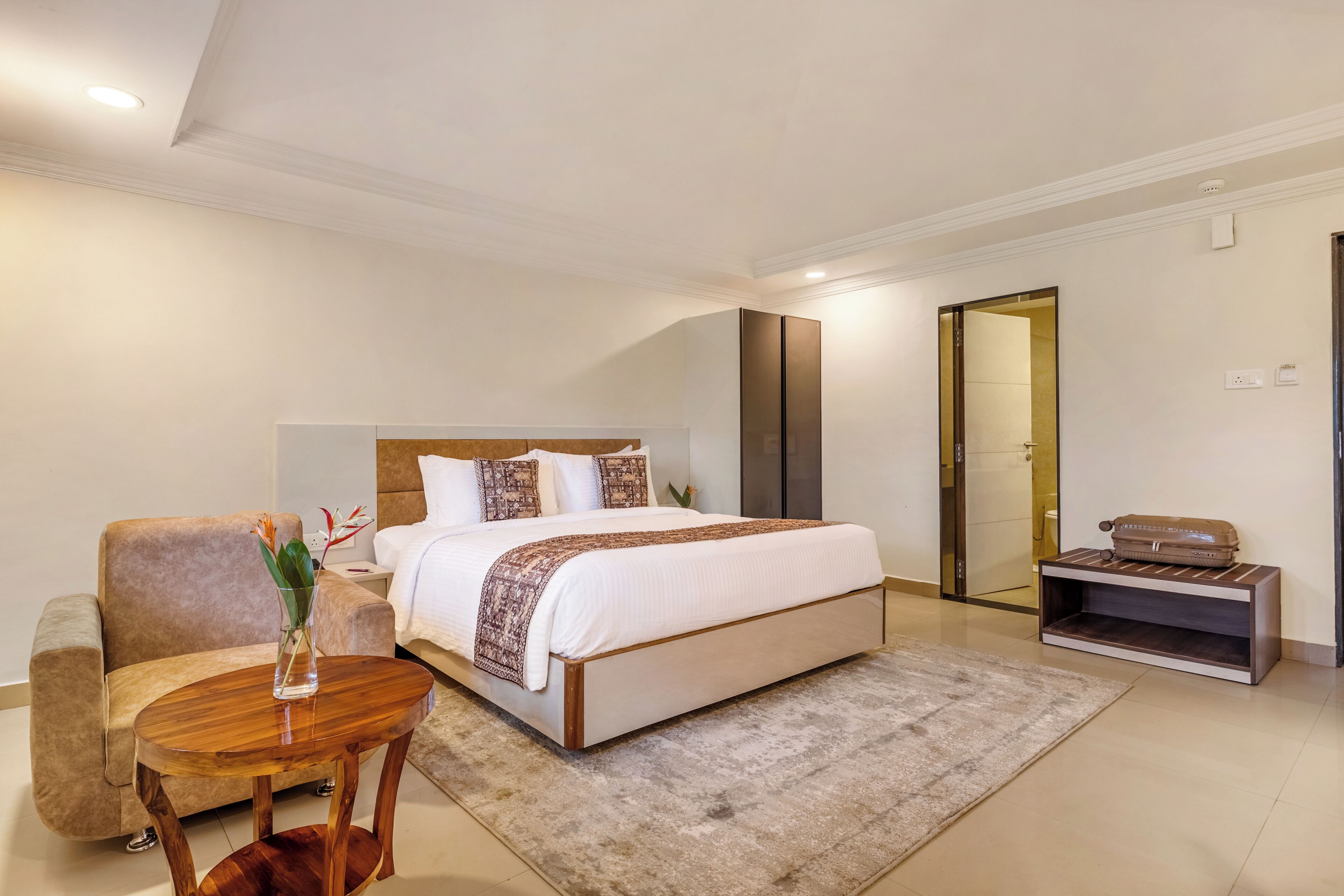 Quarto de hotel moderno com uma cama grande, área de estar com mesa e poltrona, uma pequena escrivaninha, e um banheiro visível por uma porta aberta.