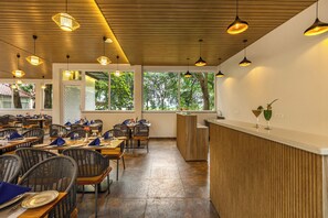 A restaurant interior with wooden ceiling, tables with chairs, and large windows with a view of trees.