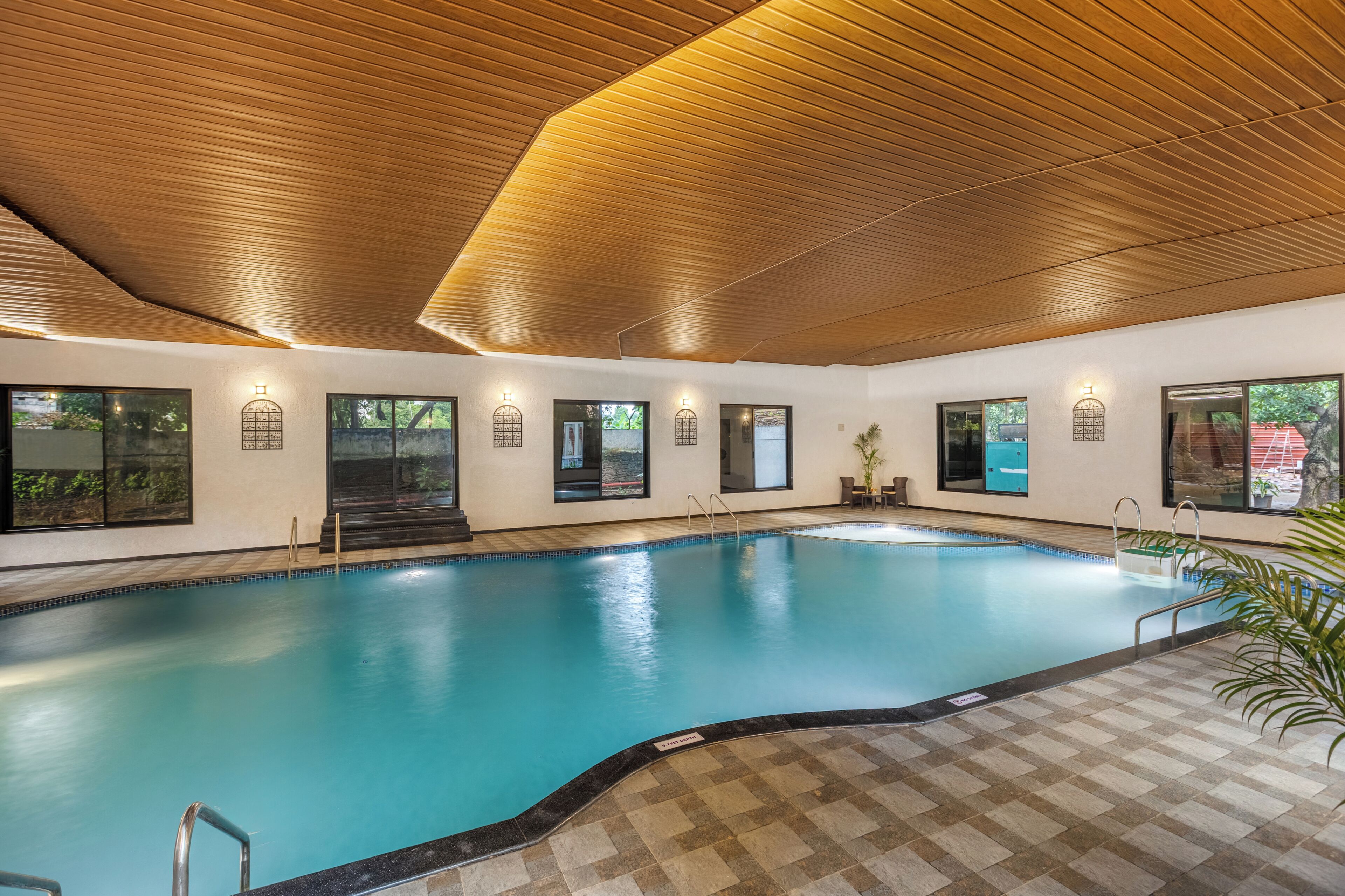 An indoor swimming pool with a wooden ceiling, clear glass windows, and a tiled floor.