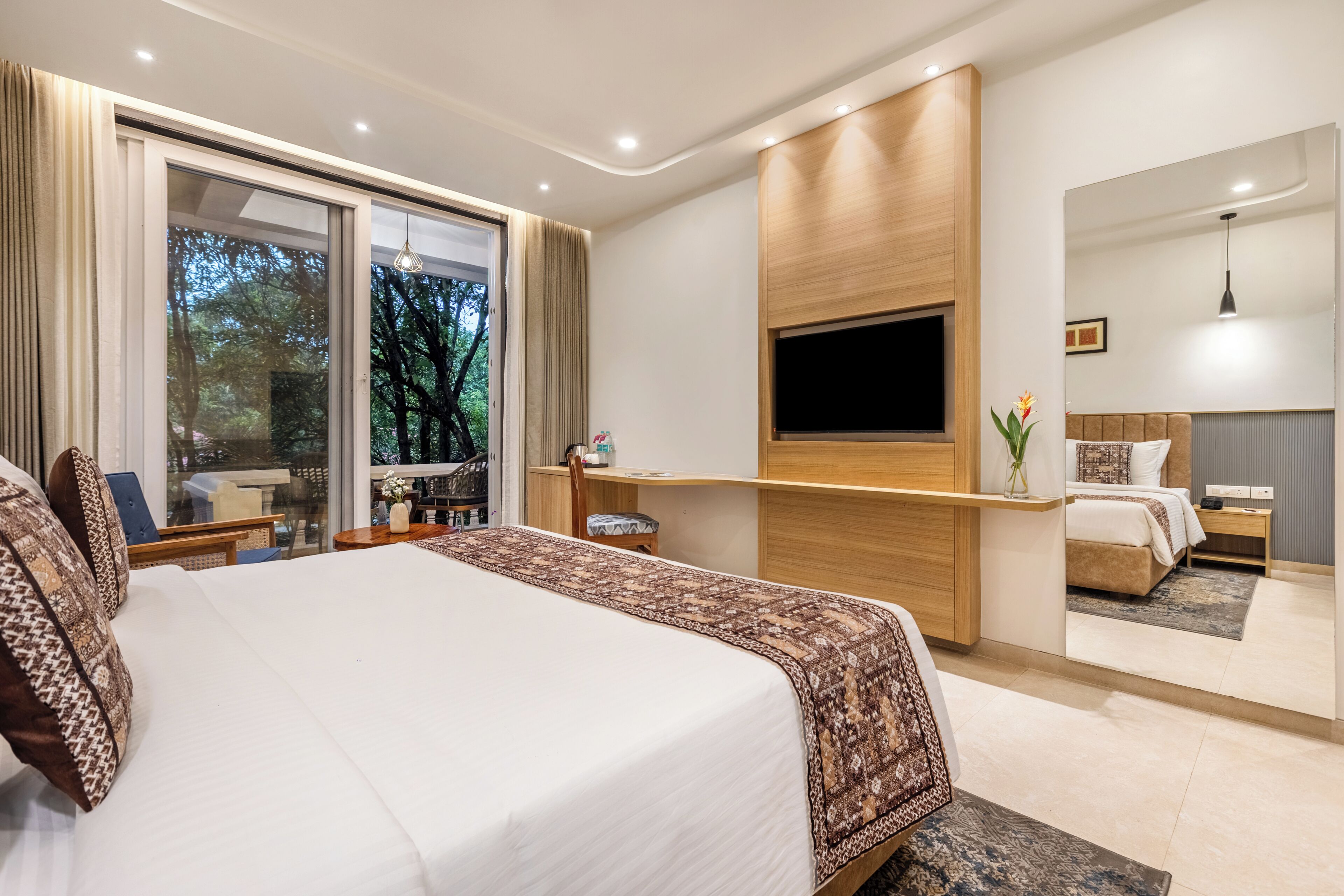 A modern hotel room with a large bed, a TV mounted on the wall, and a balcony with a view of trees.
