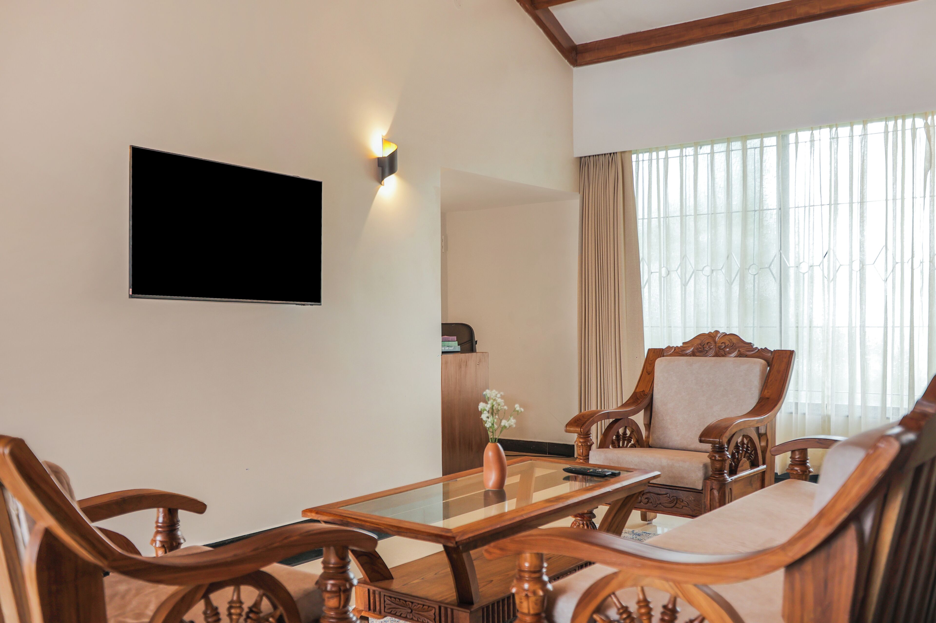A room with a wooden coffee table, two upholstered chairs, a television mounted on the wall, and a window with curtains.