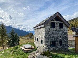 A stone building with a sloped roof, a circular outdoor seating area, and a mountainous backdrop. - AffittiPontediLegno - Chalet Baita Aria (Passo del Tonale)