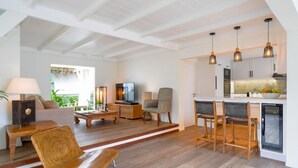 Una sala de estar moderna con un sofá, una mesa de centro y una televisión. Hay una zona de cocina con un comedor y luces colgantes.