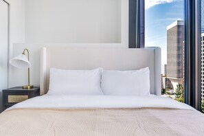 Ein Hotelzimmer mit einem großen Bett, einem Nachttisch mit Lampe und Blick auf hohe Gebäude durch ein Fenster.