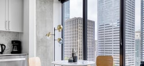 A modern kitchen with a coffee maker, a round table with a vase, and a view of tall buildings through large windows. - Blueground | First Hill, rooftop pool, nr Seattle Art Museum (Seattle)