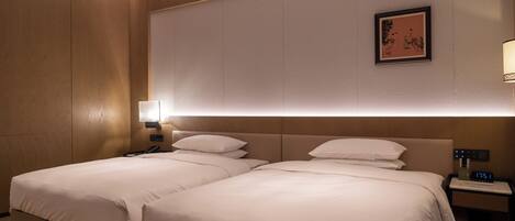 A hotel room with two beds, a bedside table with a lamp, a wall-mounted artwork, and a digital clock.