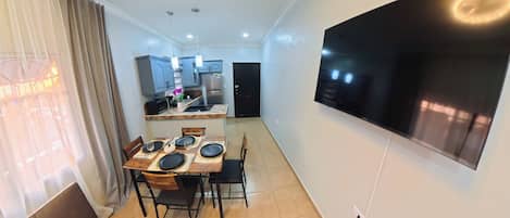 A dining area with a table set for four, a flat-screen TV mounted on the wall, and a kitchen in the background.