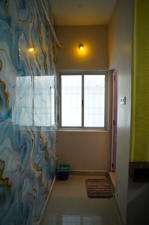 Una habitación con ventana, una pared con diseño de mármol, un cubo de basura verde y un felpudo marrón en el suelo.