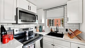 Une cuisine avec des meubles blancs, un four à micro-ondes en acier inoxydable, une cuisinière noire et une fenêtre avec des stores.