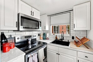 Una cocina con alacenas blancas, un microondas de acero inoxidable, una estufa negra y una ventana con persianas.