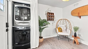 Une buanderie équipée d’un lave-linge et d’un sèche-linge, d’une chaise en osier, d’une petite table en bois et d’une planche de surf accrochée au mur.