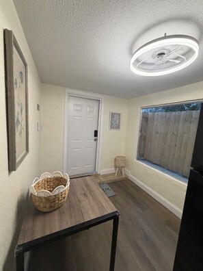 A room with a wooden floor, a white door, a ceiling fan, a window with a view, and a basket on a table. - Loft Living: 4 to 5 minutes from Bush Gardens (Tampa)