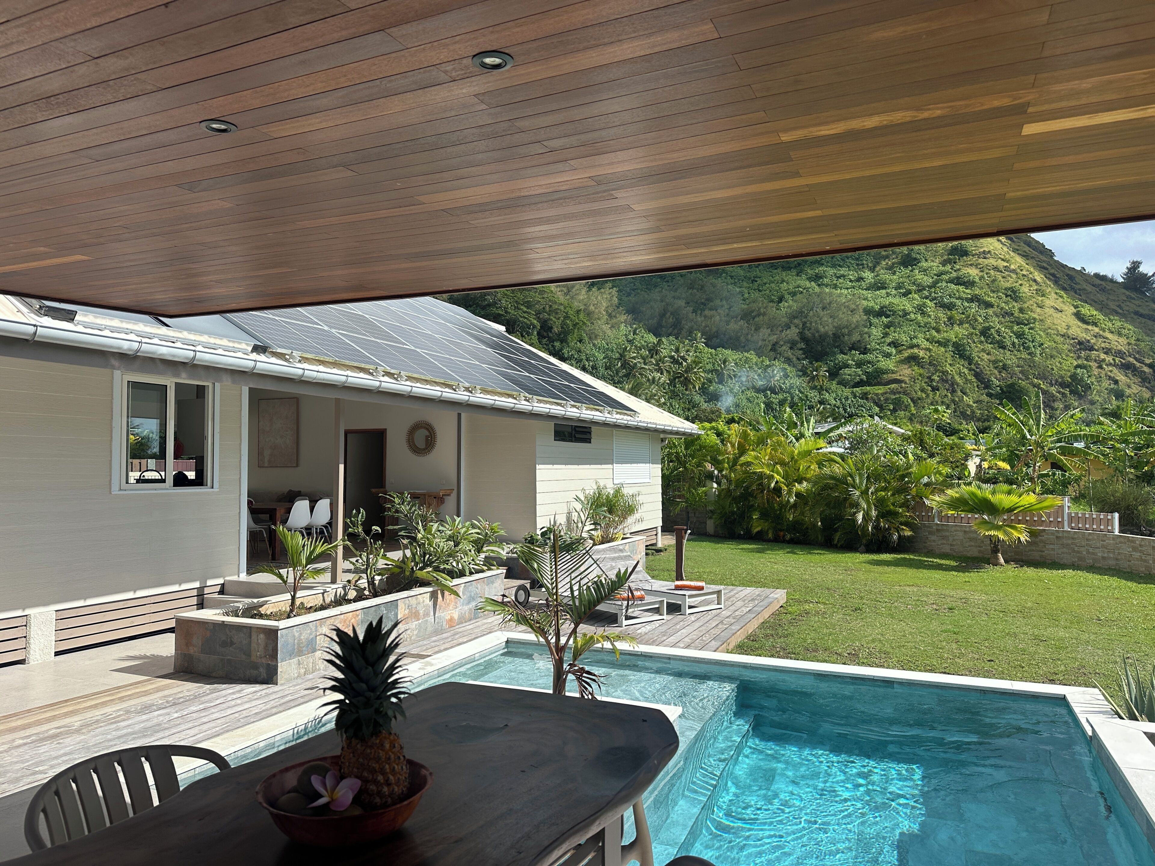 A poolside area with a wooden deck, a table with a pineapple centerpiece, and a view of a house with solar panels and a lush green landscape.