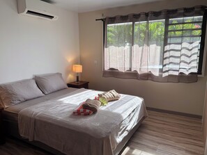 A neatly made bed with a checkered blanket, a bedside lamp, and a window with a view of greenery. - Villa Noa Vai • Swimming Pool & Lagoon Access by Kayak (papetoai)