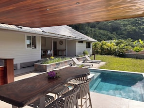 A poolside area with a wooden table and chairs, a covered patio, and a view of a lush garden and hills. - Villa Noa Vai • Swimming Pool & Lagoon Access by Kayak (papetoai)