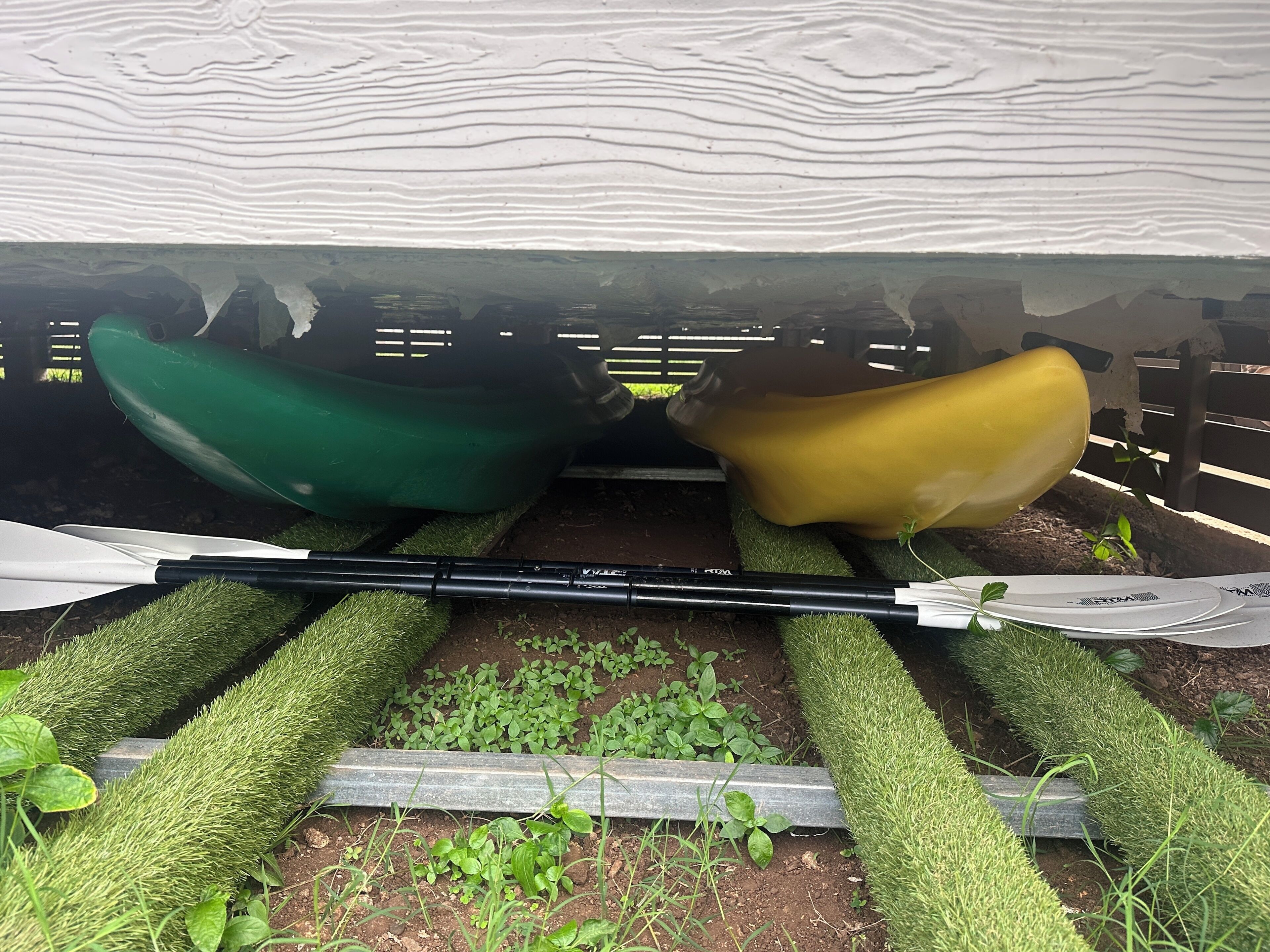Caiaque verde e amarelo apoiado em superfície gramada, com um cano de metal e um remo branco na frente.
