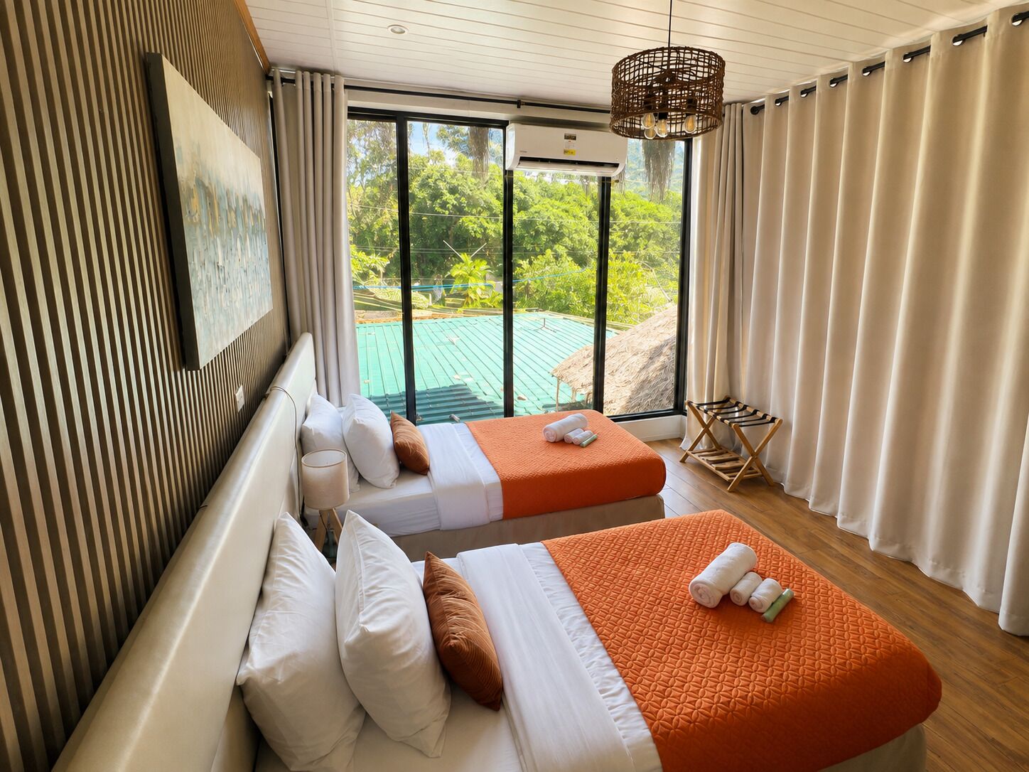 A hotel room with two beds, a large window with a view of greenery, and a hanging light fixture.