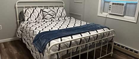 A single bed with a metal frame, a patterned bedspread, and a blue blanket. There is a window with an air conditioning unit on the wall.