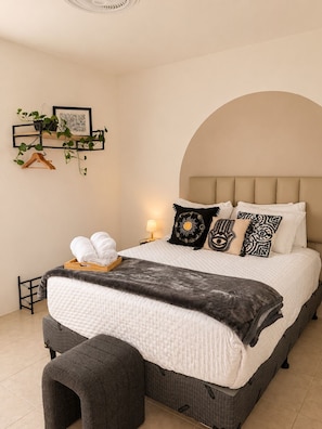 A neatly made bed with a headboard, a bedside table with a lamp, and a wall-mounted shelf with plants and a picture.