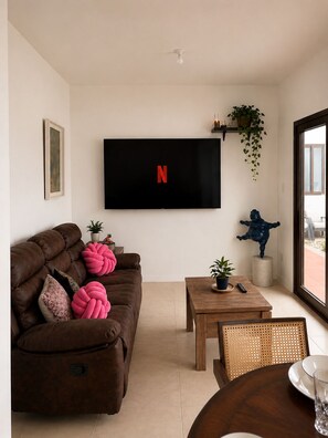A living room with a brown sofa, a coffee table, a TV mounted on the wall, and a window with a view.