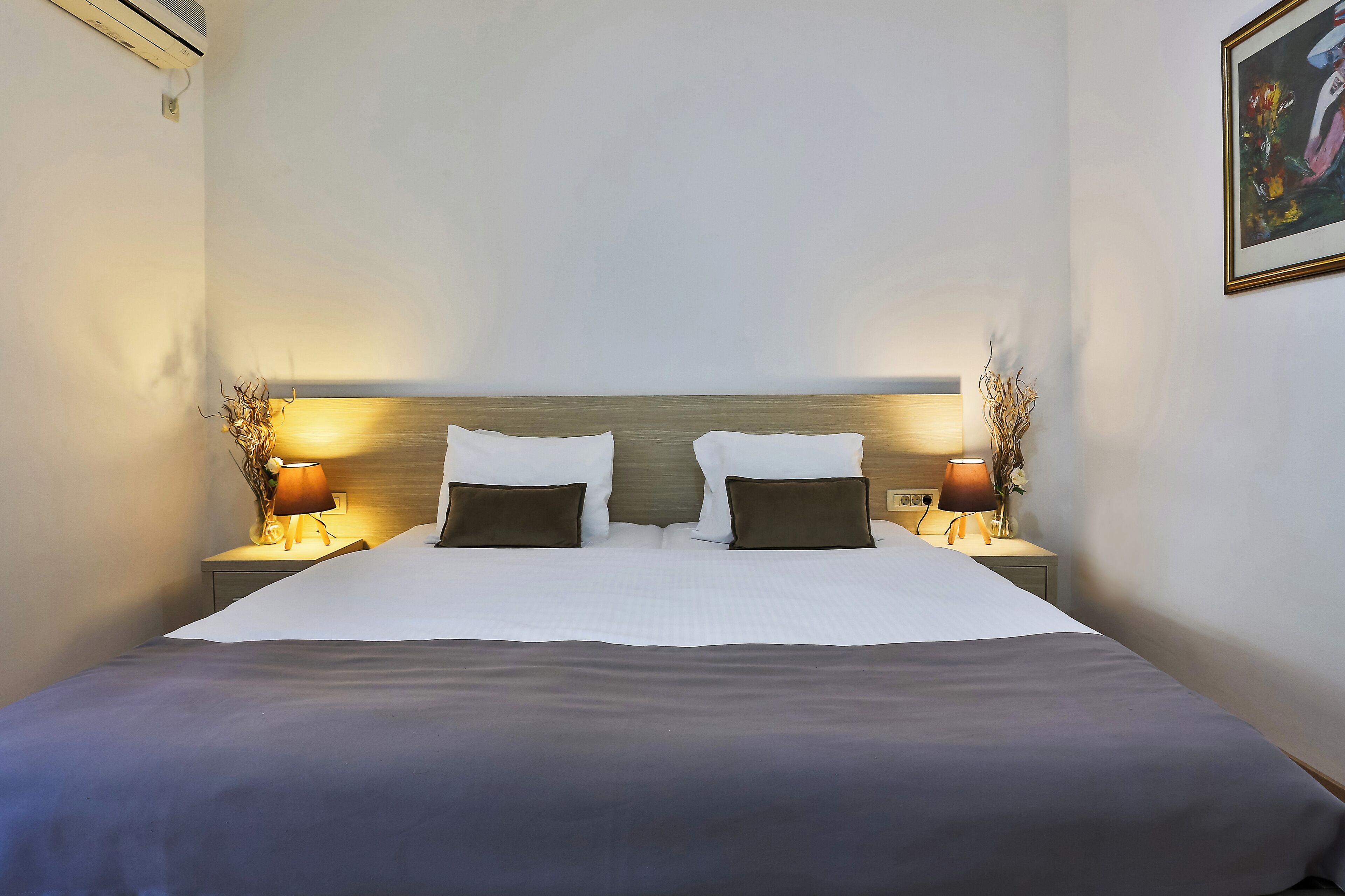 Una cama bien hecha con ropa de cama blanca y marrón oscuro, dos lámparas de noche y una obra enmarcada en la pared.