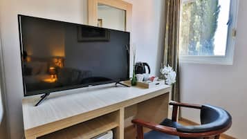 A modern room with a large flat-screen TV on a wooden desk, a chair, a small table with a vase of flowers, and a window with a view of trees.
