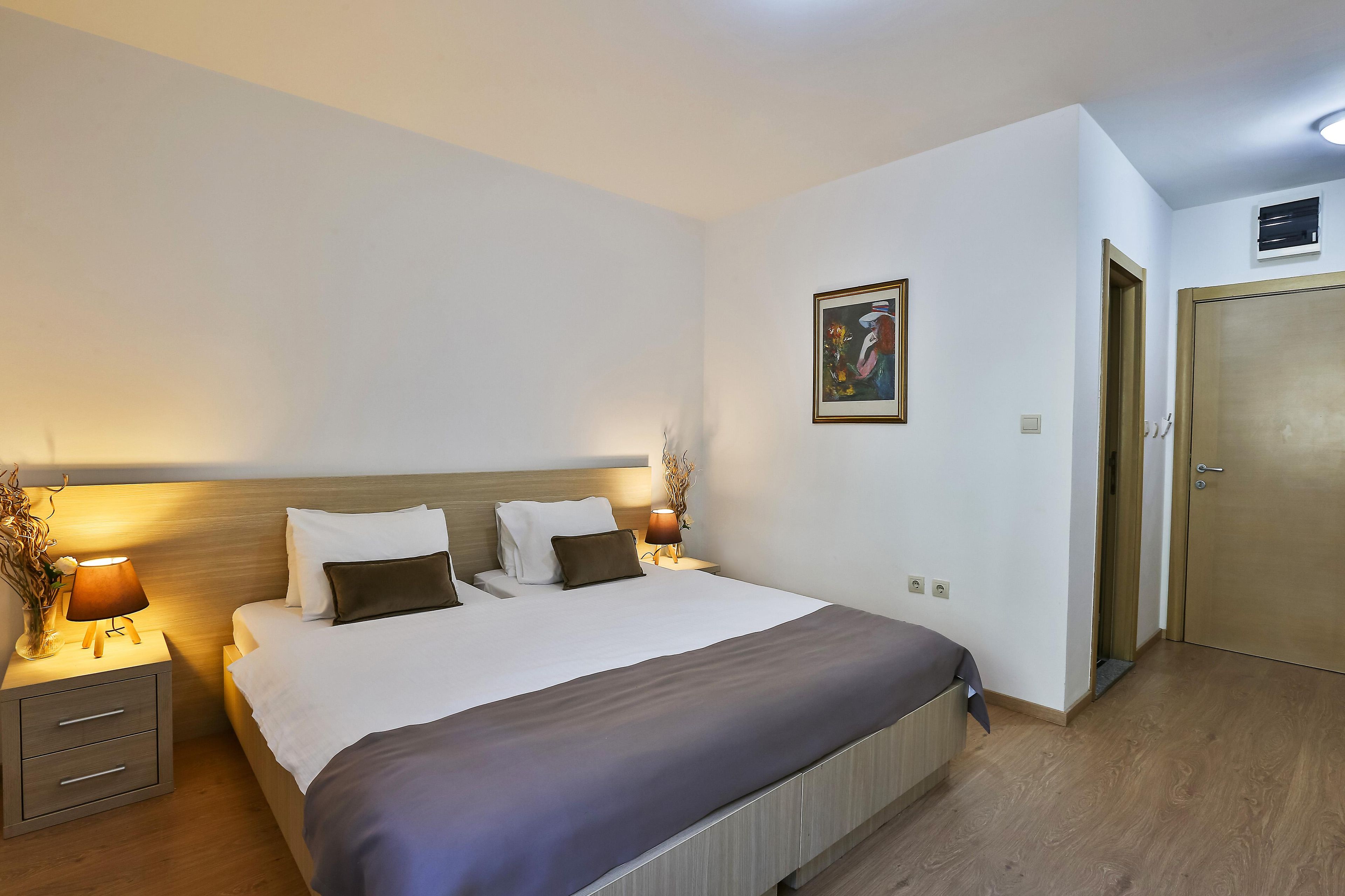 Una habitación de hotel con una cama, lámparas de noche, un cuadro en la pared y una puerta.