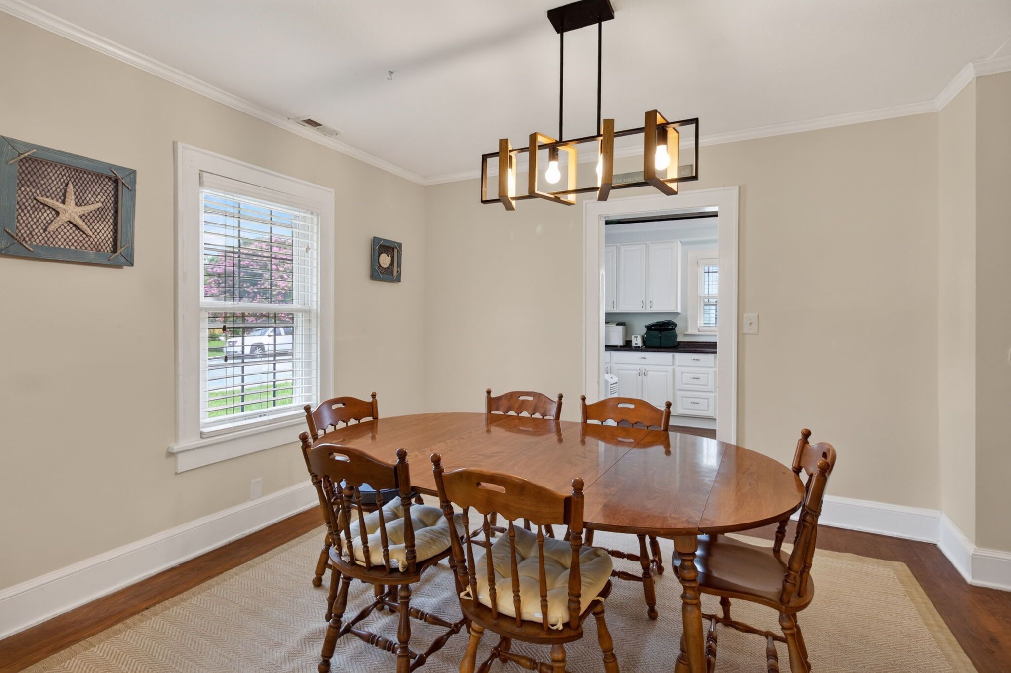 Uma sala de jantar com uma mesa redonda de madeira, seis cadeiras, um candelabro, uma janela com persianas e uma cozinha visível através de uma porta aberta.