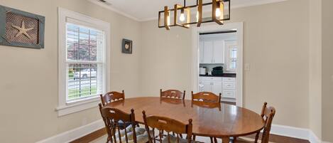 Uma sala de jantar com uma mesa redonda de madeira, seis cadeiras, um candelabro, uma janela com persianas e uma cozinha visível através de uma porta aberta.