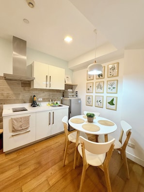 A modern kitchen with white cabinets, a stainless steel range hood, and a built-in oven. There is a dining area with a white table and chairs, and a wall decorated with framed botanical prints. - Modern 1BR Staycation in Acqua Private Residences (Mandaluyong City)
