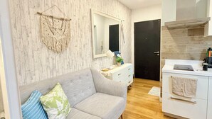 Ein modernes Wohnzimmer mit grauem Sofa, Wandbehängen aus Makramee, einem großen Spiegel und einer Küchenzeile mit Herd und Schränken.