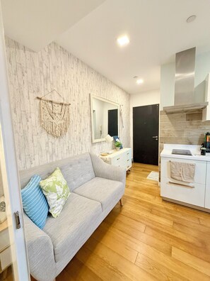 A modern living room with a grey sofa, a macrame wall hanging, a large mirror, and a kitchen area with a stove and cabinets. - Modern 1BR Staycation in Acqua Private Residences (Mandaluyong City)