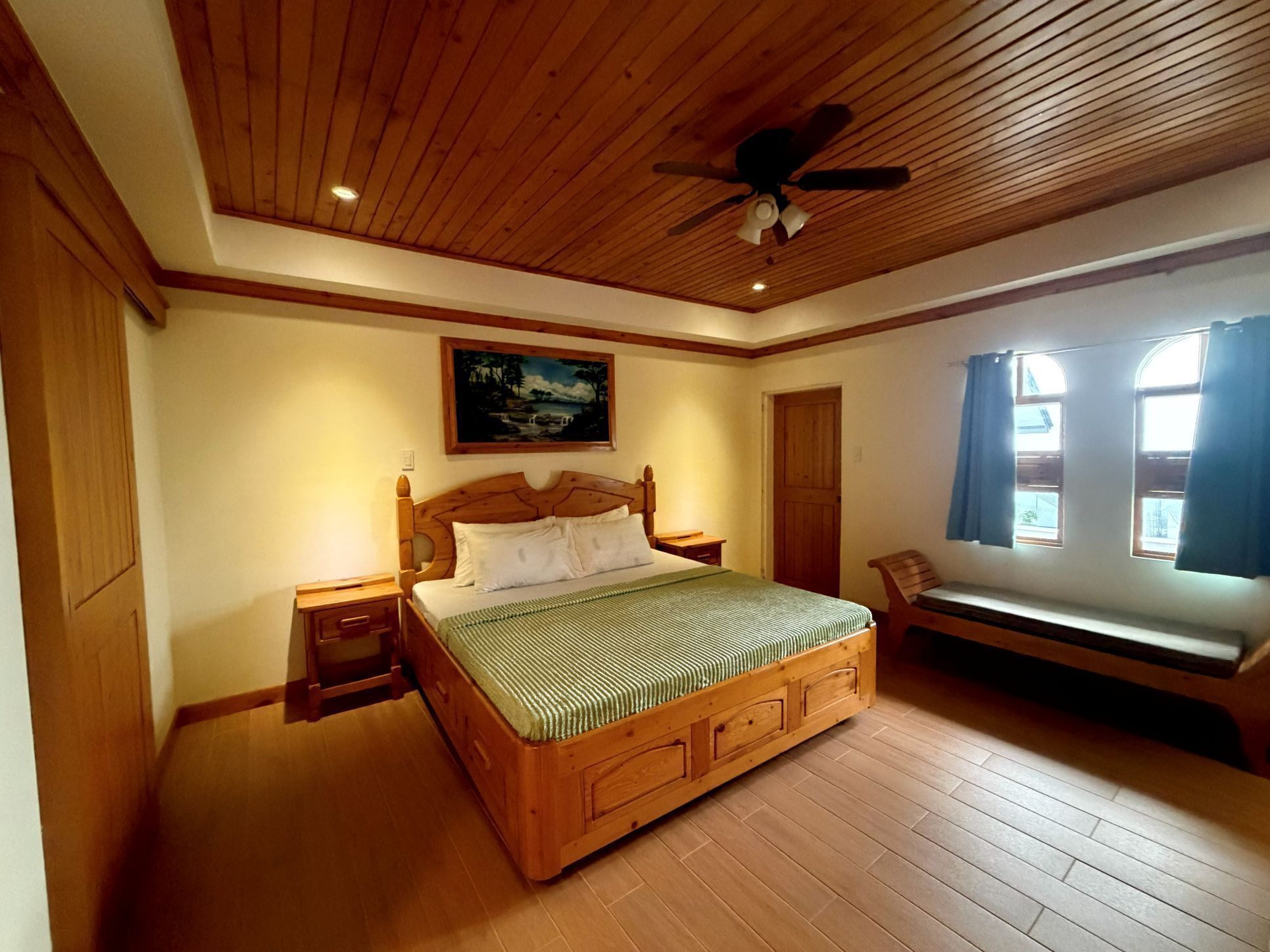 Une chambre à coucher avec un lit en bois, une table de chevet, un banc, un ventilateur de plafond et un tableau accroché au mur.