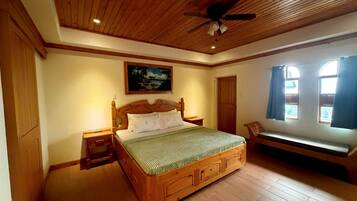 Une chambre à coucher avec un lit en bois, une table de chevet, un banc, un ventilateur de plafond et un tableau accroché au mur.