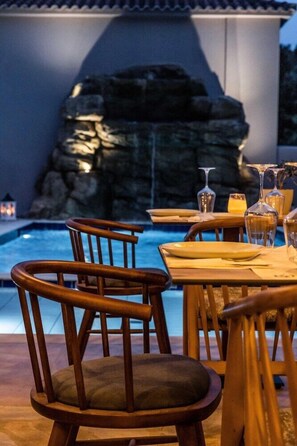 Un área de comedor junto a la piscina con sillas y mesas de madera, una pared de piedra y una piscina.