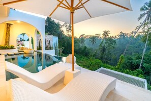 Uma área de piscina moderna ao ar livre, com espreguiçadeiras e um grande guarda-sol, rodeada por vegetação tropical exuberante.