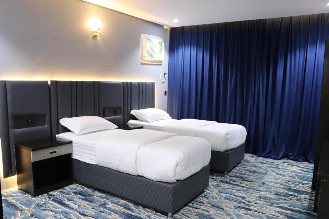 Quarto de hotel com duas camas, cabeceira, mesas de cabeceira, ar condicionado fixado na parede e cortina azul.