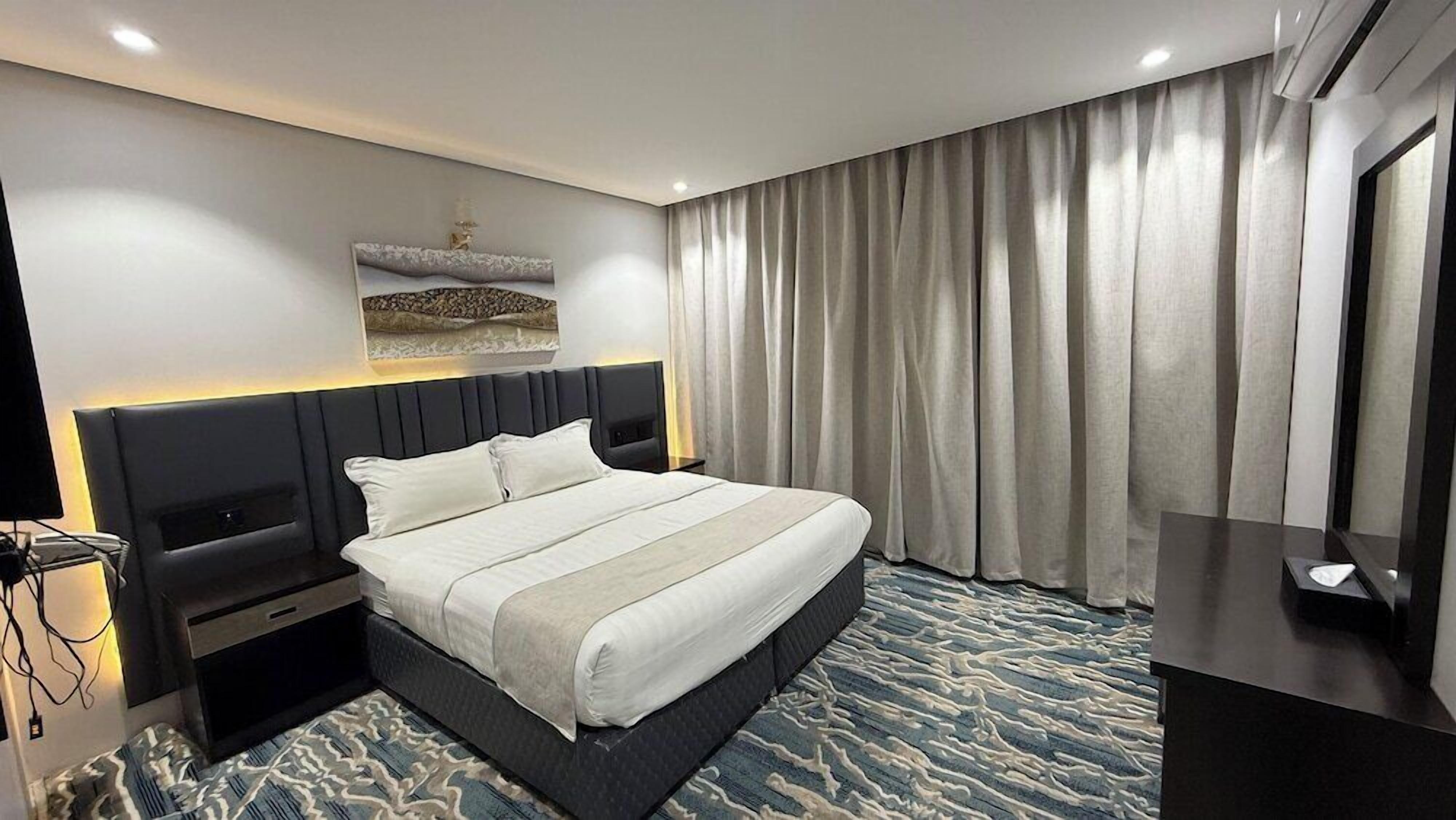 Habitación de hotel con cama, mesitas de noche, televisor de pantalla plana y un amplio ventanal con cortinas.