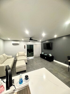 A modern living room with a flat-screen TV mounted on the wall, a ceiling fan, and a seating area with a sofa and ottoman. - Beachfront Paradise!
1 Bedroom First Floor Condo (Cocoa Beach)