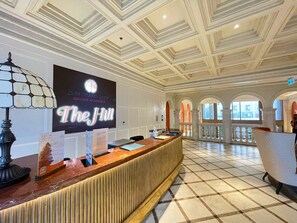 Uma área de recepção com balcão de check-in, uma placa grande e iluminada e piso de azulejos.