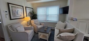 Uma sala de estar com dois poltronas, uma mesa de centro, uma luminária, uma televisão e quadros nas paredes.