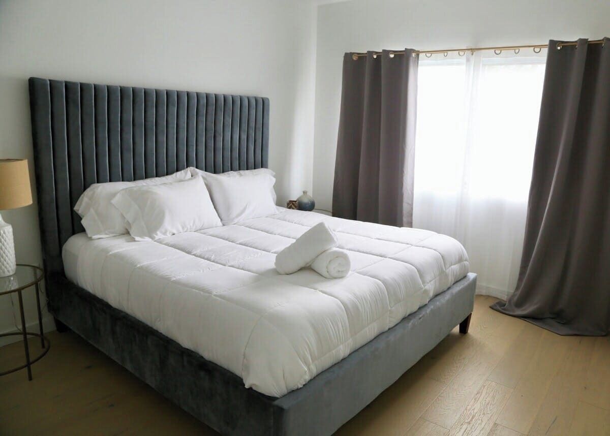 A neatly made bed with white linens and a folded towel, a bedside table with a lamp, and a window with curtains.