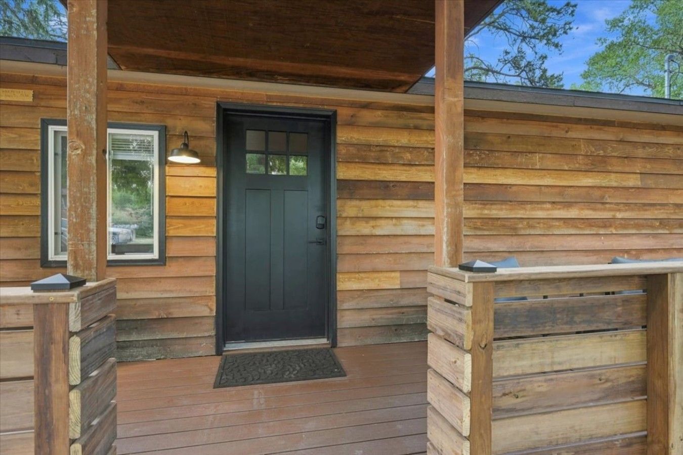 A wooden cabin with a black door, a porch with a bench, and a window with a light fixture.