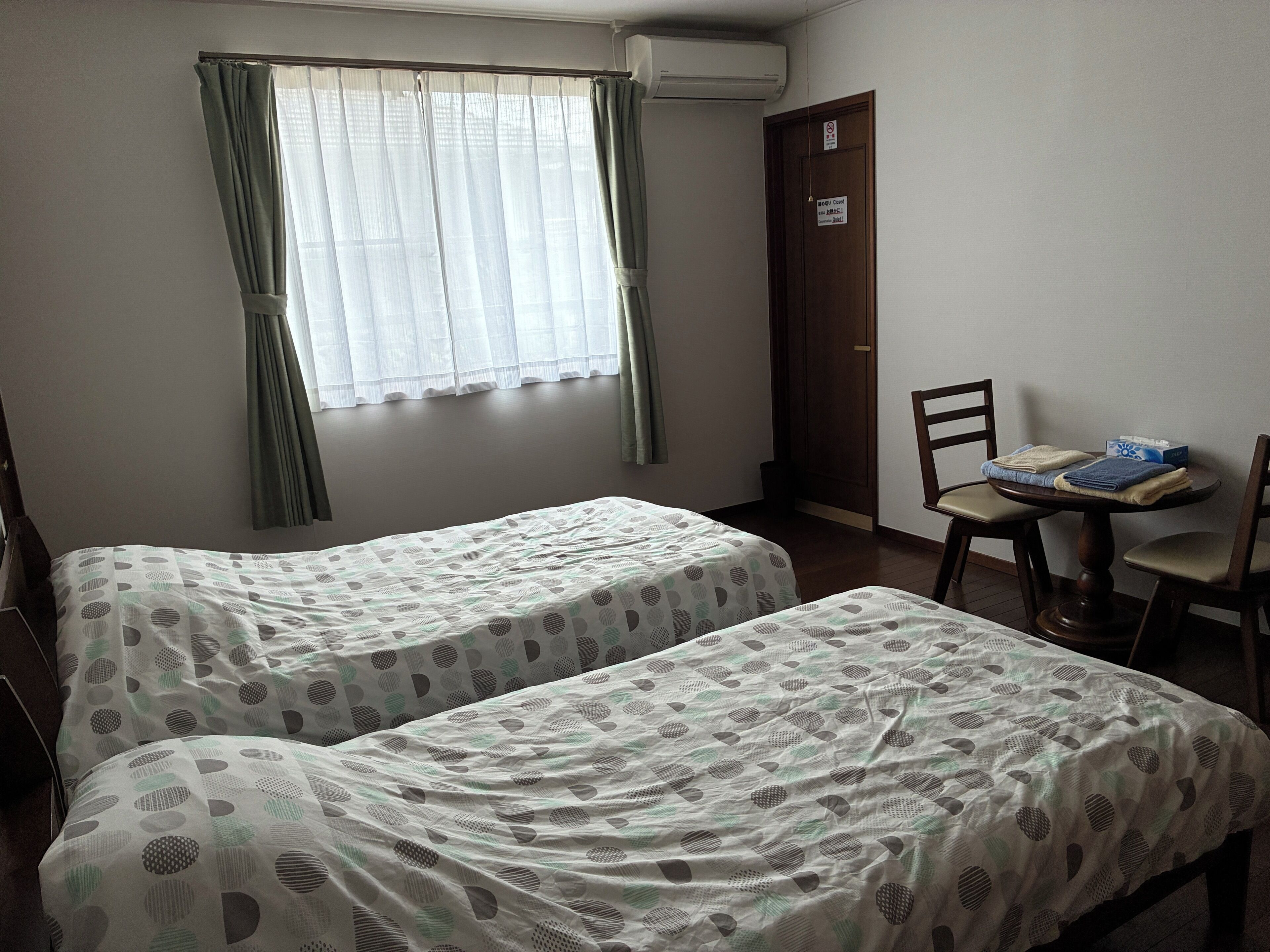 A room with two beds, a table with chairs, a window with curtains, and a door with a sign.
